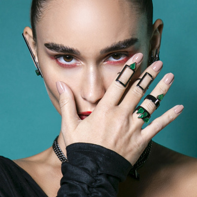 Model with bold makeup and various jewelry pieces, including rings and earrings, posing with hand on face.