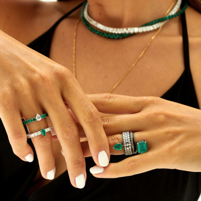 Close-up of hands displaying various emerald and diamond rings and a necklace.