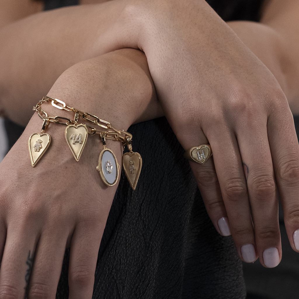 Gold charm bracelet with various pendants and a heart-shaped ring on a woman's hand, showcasing elegant jewelry design.