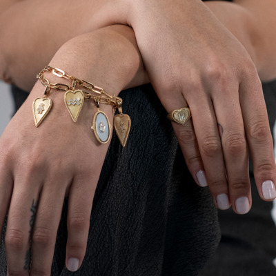 Gold charm bracelet with various pendants and a matching gold ring worn on hands.