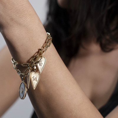 Gold charm bracelet featuring heart and oval pendants on a person's wrist.