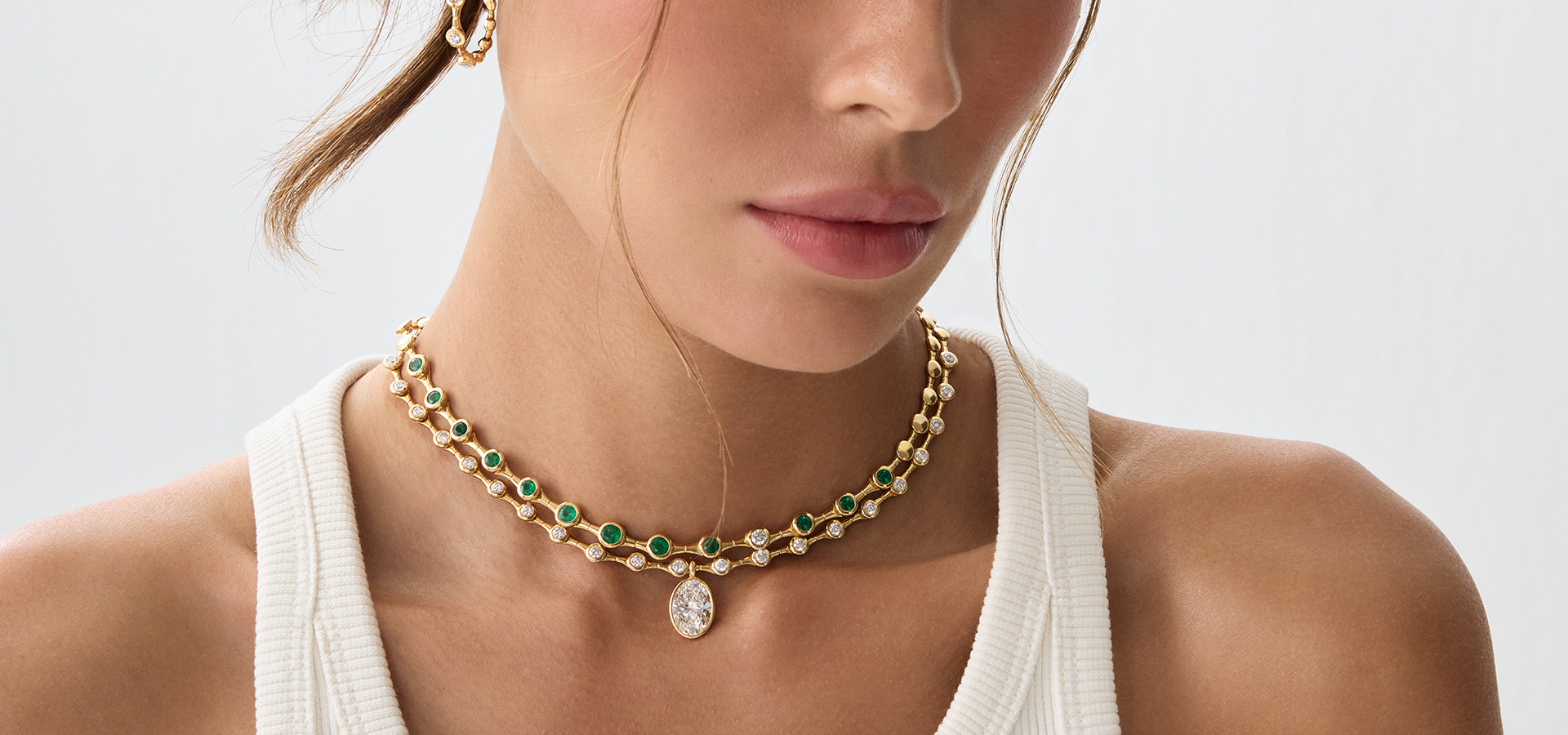 Close-up of a model wearing a gold two-strand necklace with alternating green gemstones and diamonds, paired with a white ribbed tank top